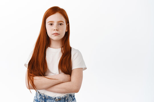 Serious Little Girl With Ginger Hair Cross Arms On Chest And Look Determined. Cofident Redhead Kid With Freckles Staring Focused At Camera, White Background