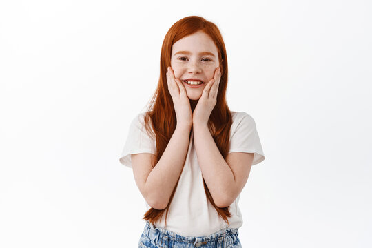 Cute little girl with long red hair and freckles, touch her face and smile, look surprised, receive a gift and rejoice. Redhead child with adorable grin staring at camera, white background