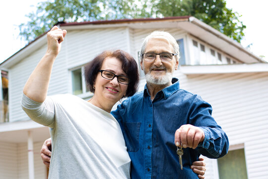 Smiling Senior Couple Holding Keys To New Own Housing, Mature Family Bonding To Each Other And Waving Hand Looking At Camera, Standing In Front Of Big White House.Happy And Prosperous Old Age Concept