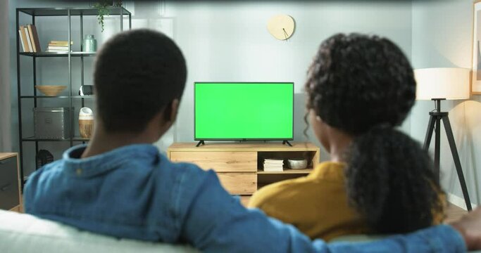 Close up of wife and husband sitting at home together watching TV with chroma key. Back view of African American couple man and woman sitting on sofa watch television program, green screen