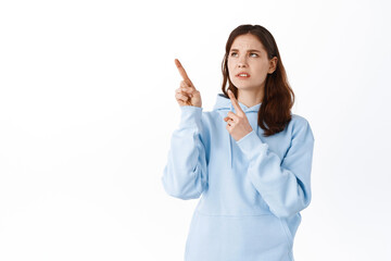 Girl cringe from something disgusting. Displeased young woman pointing and looking at upper left corner with aversion and dislike, grimacing, standing over white background