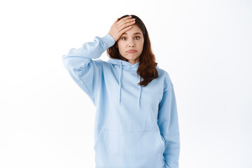 Troubled young woman forgot or remember something, holding hand on forehead, making facepalm and pouting bothered, having problem, standing against white background