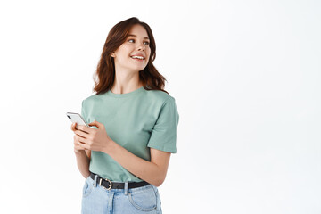 Young modern girl standing with smartphone and look aside at copy space, smiling while chatting on social media app, standing against white background