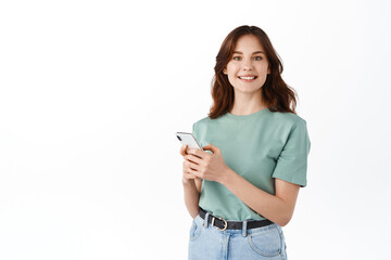 Attractive woman in t-shirt using smartphone messanger, chatting on phone and smiling at camera, reading social media, standing against white background