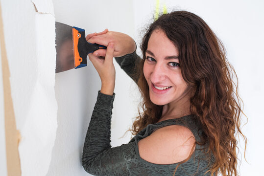 Smiling woman holding a scraper while removing wallpaper from a wall. Home renovations.
