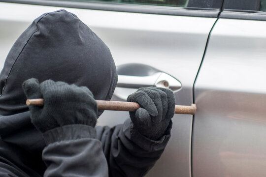 Robber Man In Black Hoodie Jacket Using A Crowbar To Break Lock And Steal A Vehicle. Car Thief Or Theft For Insurance Concept