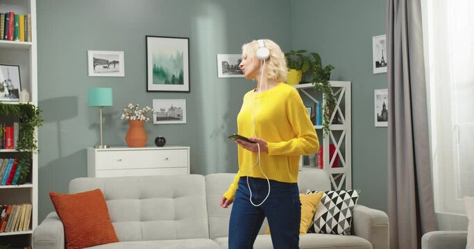 Portrait of Caucasian happy beautiful blonde woman dancing having fun in modern living room in apartment while listening to fav song in headphones from smartphone. Leisure, relax concept