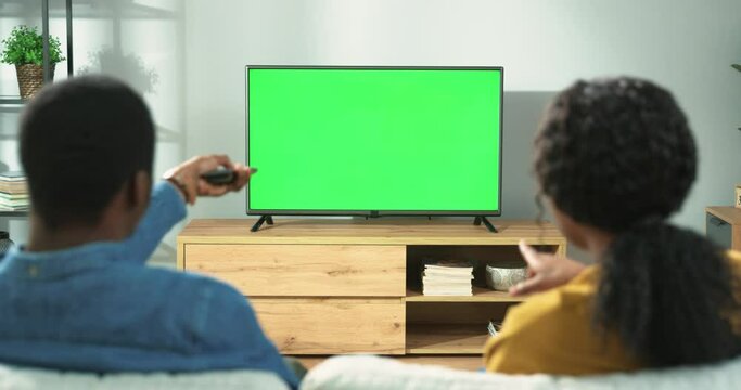 Back view of young African American family couple watching TV with chroma key while resting at home sitting on sofa in room. Wife and husband spending time together watch television with green screen