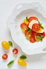 Caprese. Fresh colorful tomatoes and basil leaves in a ceramic bowl on a white background. Home made food. Concept for a tasty and healthy appetizer, flat lay. 