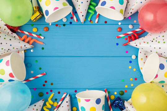 Top view photo of birthday party composition spiral candles pipes striped straws hats confetti balloons polka dots paper cups and plates on isolated blue wooden table background with copyspace