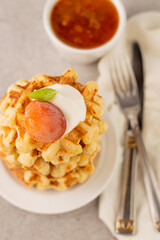 Blurred image of Belgian waffles with apricot and cream in a white plate, knife, fork, gravy boat with apricot jam on a gray background.