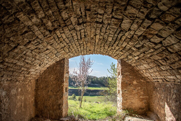 old stone building with arched roof opening to the outside where you can see a flowering almond tree and fields.