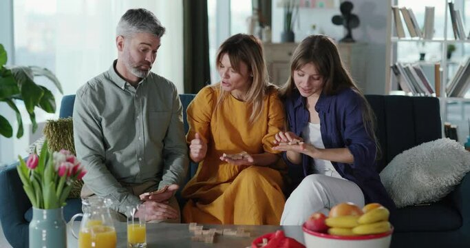 Caucasian cheerful family trio playing domino table game in cozy living room apart,ment. FRelationship. Parents with daughter. Leisure activity.