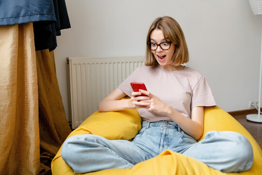 young shocked woman staring at mobile in the apartment wearing blue jeans pink tshirt and glasses