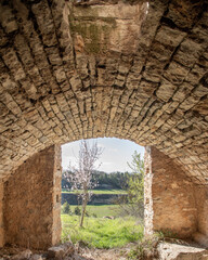 Obraz premium old stone building with arched roof opening to the outside where you can see a flowering almond tree and fields.