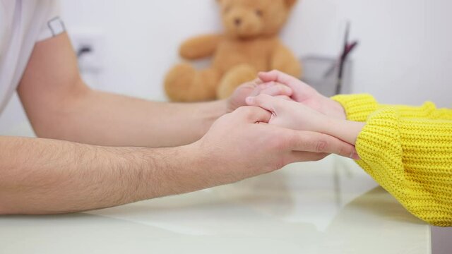 Close-up Of Male Hands Holding Child Palms With Blurred Teddy Bear At Background. Unrecognizable Middle Eastern Doctor Calming Down Girl On Appointment. Pediatric Care And Medicine.