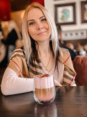 A blonde girl with a scarf on her shoulders in a soft pink sweater sits in a cozy restaurant and she is relaxed and have a coffee drink and a piece of cake on her table.