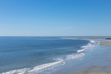 Wide flat beach on blue ocean waters, small waves and distant beachcombers, blue sky, horizontal aspect