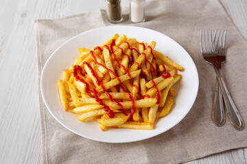 Homemade French Fries with Ketchup, Salt and Pepper on a white plate on a white wooden background, side view.