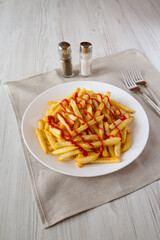 Homemade French Fries with Ketchup, Salt and Pepper on a white plate on a white wooden background, side view.
