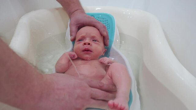 Bathing newborn baby in a baby bath, father bathing his 3-week-old son, infant in baby bath seat support.