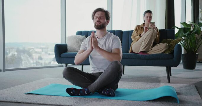 Young man doing morning exrcise training stretching muscles in lotus yoga position. His girlfriend talking mobile phone with friend on background.