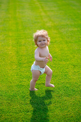 First step. Baby standing barefoot on the green lawn in diaper pants. Beautiful happy little boy...