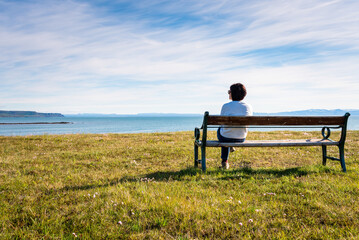 Woman sitting alone on a bench facing the ocean in Iceland on a sunny summer day. Concept of solitude.