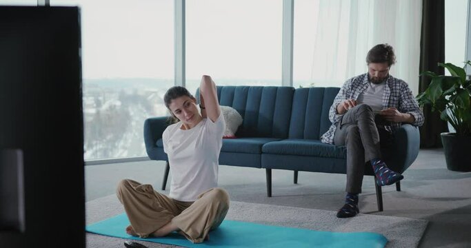 Young woman doing exercises home watching TV online workout. Her boyfriend bearded man reading story book journal on the couch.
