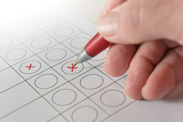 Hand holding a red ballpoint pen and makes crosses on a ballot paper, voting and election concept,...