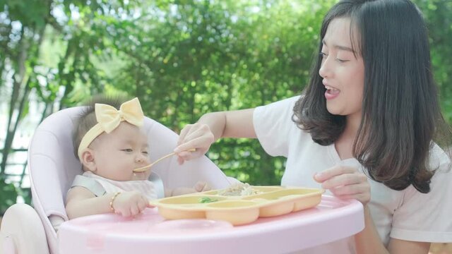 4k Mother feeding baby infant daughter toddler on highchair in a garden