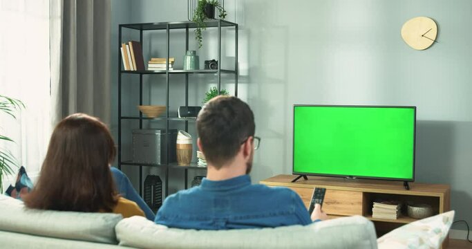Back view of young couple wife and husband watching TV with chroma key choosing program while resting at home on couch. Male and female friends watch television using remote control