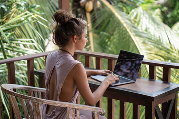 Portrait of young beautiful woman freelancer working with laptop on balcony of tropical bungalow with palm trees view