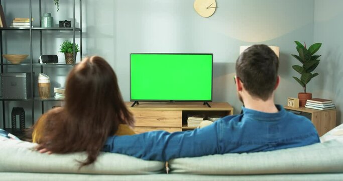 Rear of Caucasian married family young couple watching TV with chroma key and eating popcorn while sitting at home on sofa. Man and woman watch film on television in room, leisure concept
