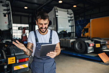Smiling tattooed bearded blue collar worker in overalls using tablet to check on delivery while...