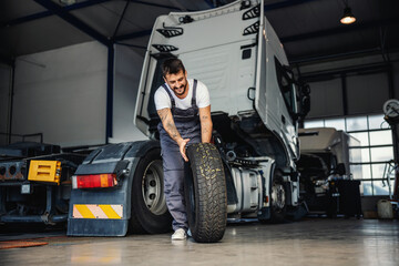 Obraz na płótnie Canvas Smiling hardworking mechanic rolling tire in order to change it on truck. He is in garage of import and export firm.