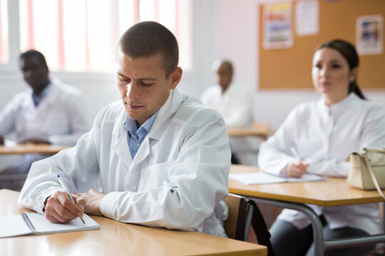 Portrait of young man in white medical coat sitting at desk in classroom attending seminar