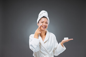 Beauty hydrating moisturizer and skincare routine. Woman in a bathrobe and a towel holds the cream and applying moisturizing cream on the cheek while standing in front of a grey background
