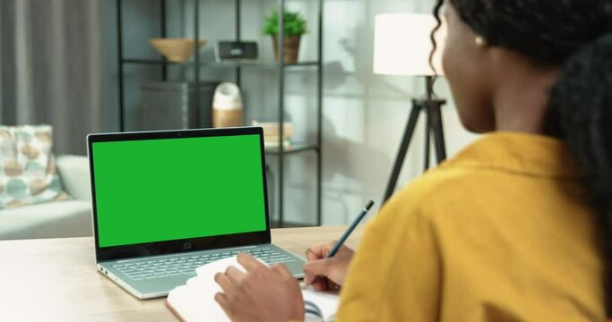 Close up Rear of African American woman student studying using laptop with chroma key while sitting at table in room in house and writing down information Browsing online on computer with green screen