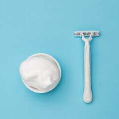 A white razor and a white bowl with shaving foam on a blue background. Flat lay.