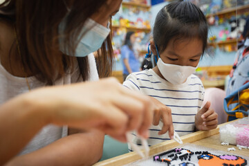 Fototapeta premium Asian child girl and her mother wearing protection mask making fusible beads thermo mosaic in pegboard that is a toy to develops the imagination of child during Coronavirus pandemic together.