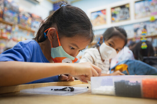 Asian child girl and sister wearing protection mask making fusible beads thermo mosaic in pegboard that is a toy to develops the imagination of child during Coronavirus pandemic.