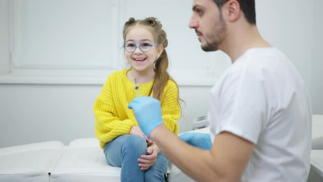 Beautiful Laughing Caucasian Girl Sitting On Medical Couch Looking At Blurred Syringe In Doctor Hands And Talking. Cute Little Patient Receiving Treatment From Middle Eastern Professional Pediatrician