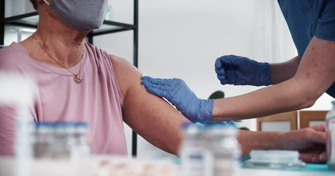 Doctor in blue scrubs, medical gloves gives vaccine injection shot with hypodermic needle to senior female patient.