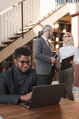 African businessman in eyeglasses sitting at the table and working on laptop with business people talking in the background