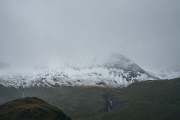 Minimalist gloomy highlands landscape with snow-capped high mountains in overcast weather. Snowbound mountain range in low clouds. Atmospheric minimalism with green mountains, white snow and gray sky.