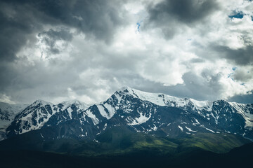 Naklejka na ściany i meble Dramatic mountains landscape with big snowy mountain ridge under cloudy sky. Dark atmospheric highland scenery with high mountain range in overcast weather. Awesome big mountains under gray clouds.