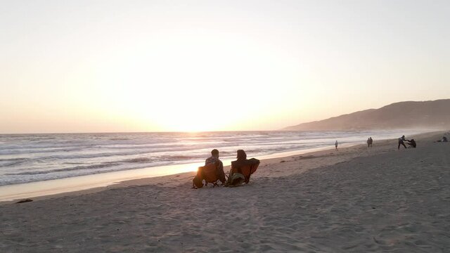 Couple In Silhouette Sitting In Folding Chairs On Beach During Sunset In Zuma Beach, Malibu, California, United States - aerial, wide shot