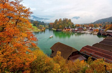 Fototapeta na wymiar A sightseeing boat cruising on Konigssee ( King's Lake ) surrounded by colorful autumn trees and boathouses on a foggy misty morning~ Beautiful scenery of Bavarian countryside in Berchtesgaden Germany