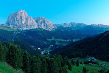 Obraz premium Scenery of majestic Sassolungo-Sassopiatto mountains at blue dusk & lights from village houses in the green grassy valley in Col Raiser, Val Gardena, Dolomiti National Park, South Tyrol, Italy, Europe